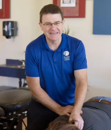 Smiling male chiropractor in blue In Line Family Chiropractic shirt adjusting a patient lying face down.