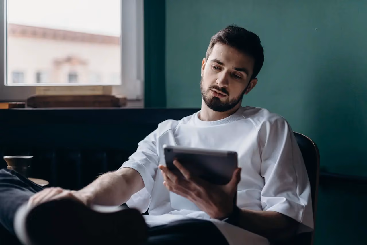 Man in white shirt sitting indoors holding a tablet and looking at its screen thoughtfully.