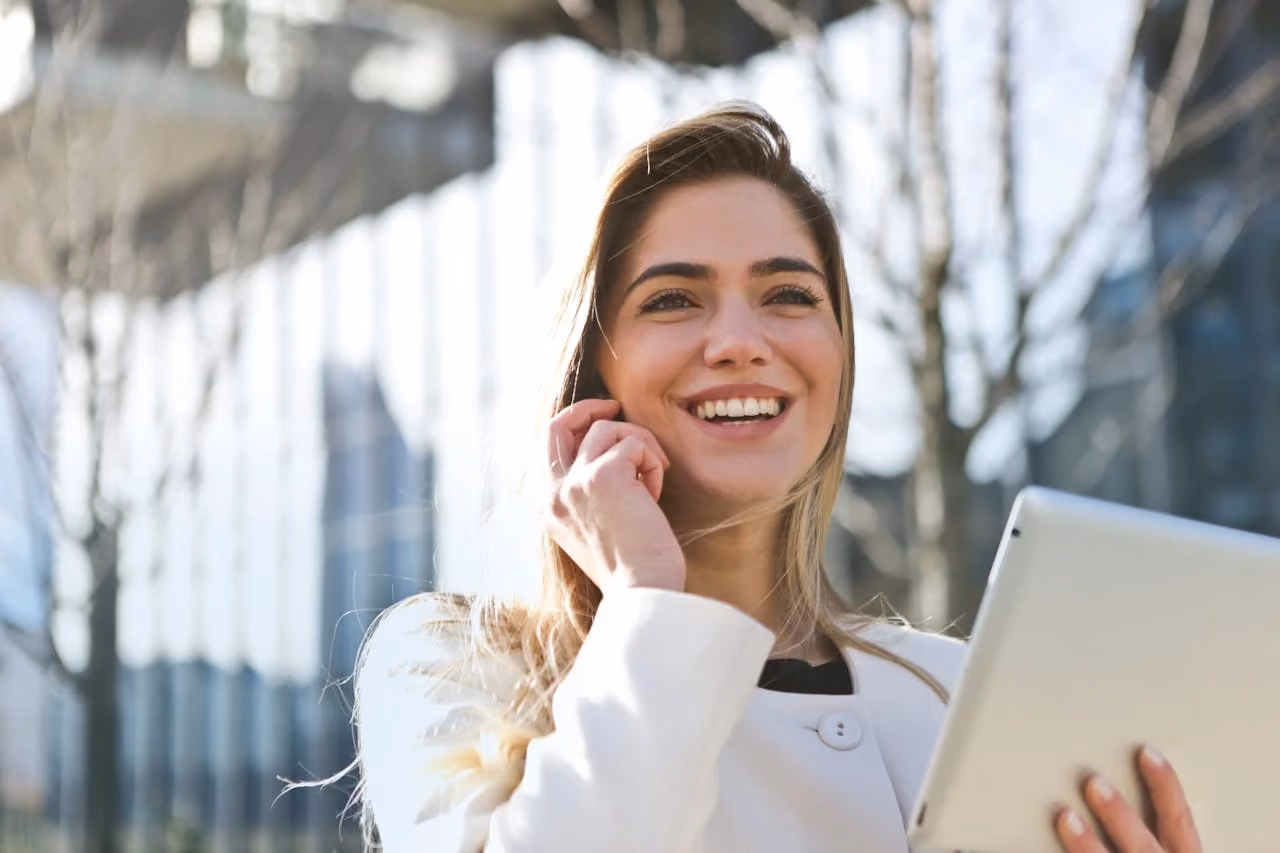 Smiling woman outdoors holding a tablet and touching her hair in daylight with blurred buildings behind.
