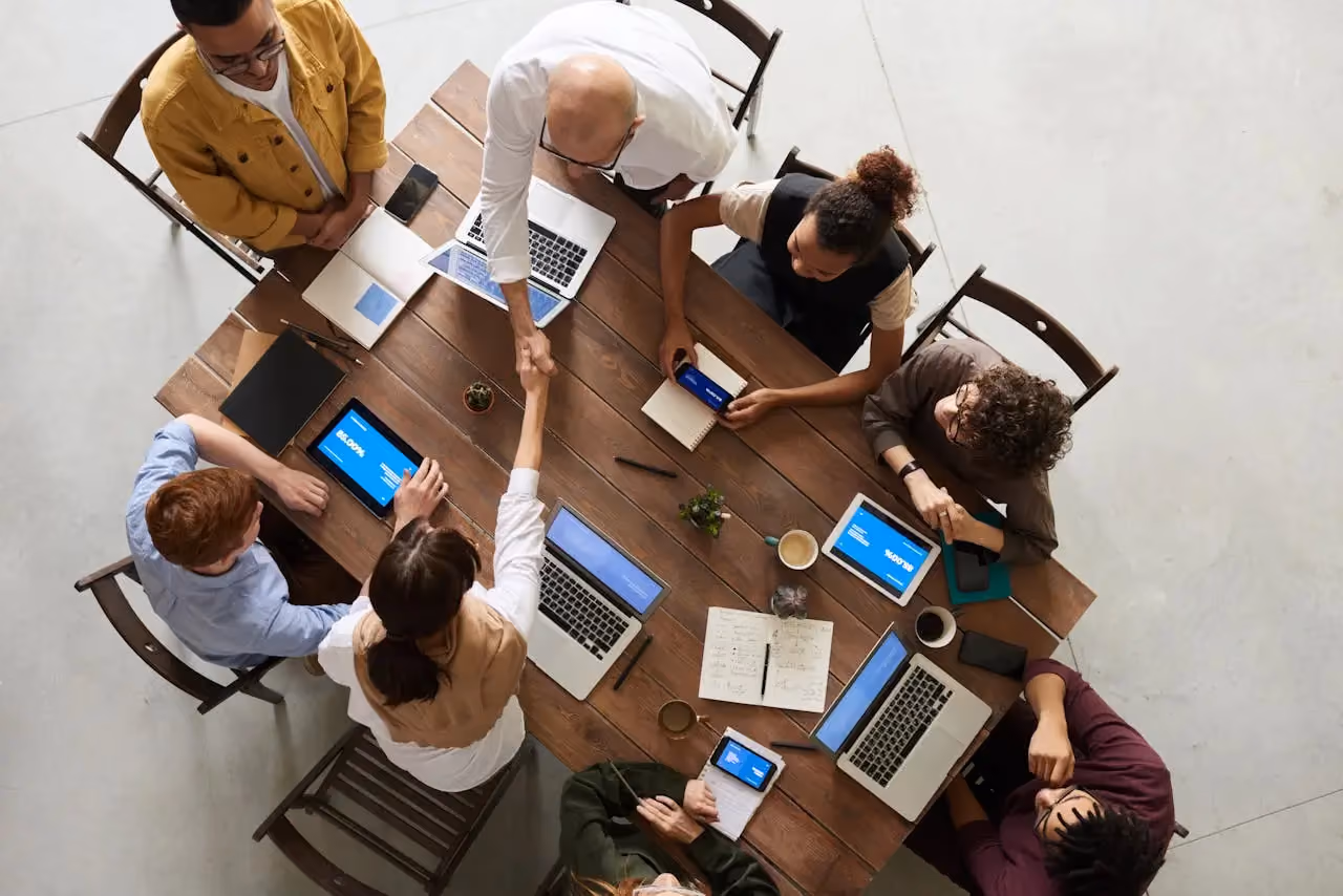 Overhead view of diverse team collaborating around a wooden table with laptops and tablets.