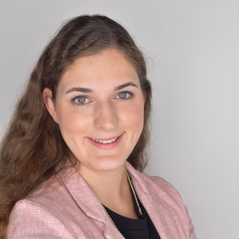 Smiling woman with long wavy hair wearing a pink blazer and black top against plain background.