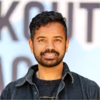 Smiling man with short curly hair and beard wearing a dark jacket in front of a blurred background.