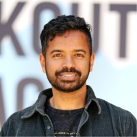 Smiling man with short curly hair and beard wearing a dark jacket in front of a blurred background.