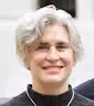 Smiling person with short, wavy gray hair wearing a dark top in a bright indoor setting.