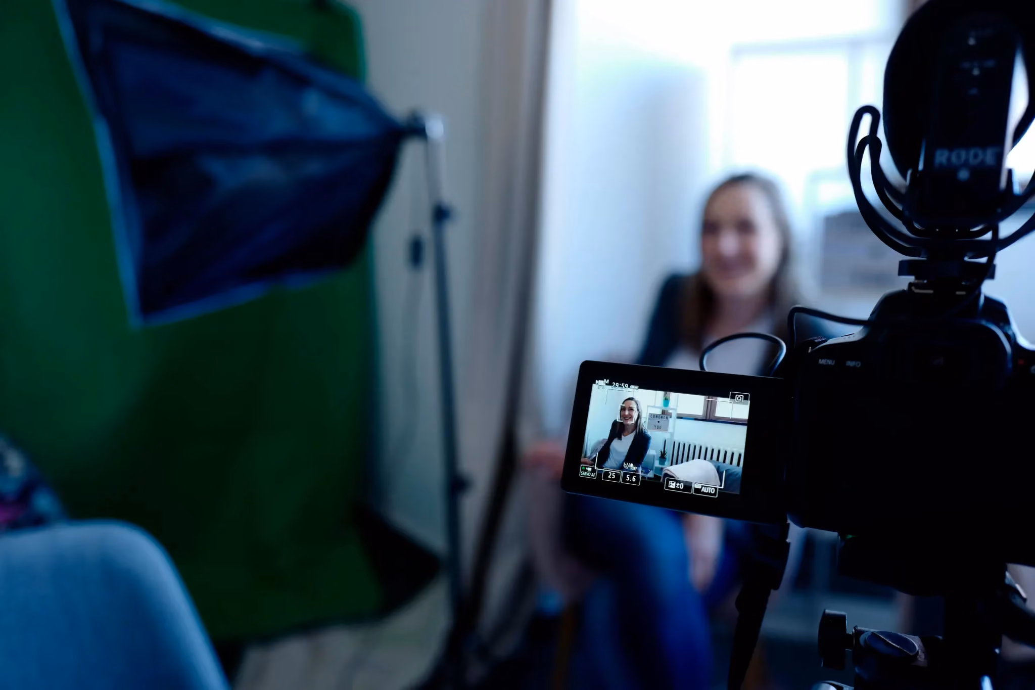 Camera screen shows a woman smiling while sitting in a recording setup with a green screen and light.