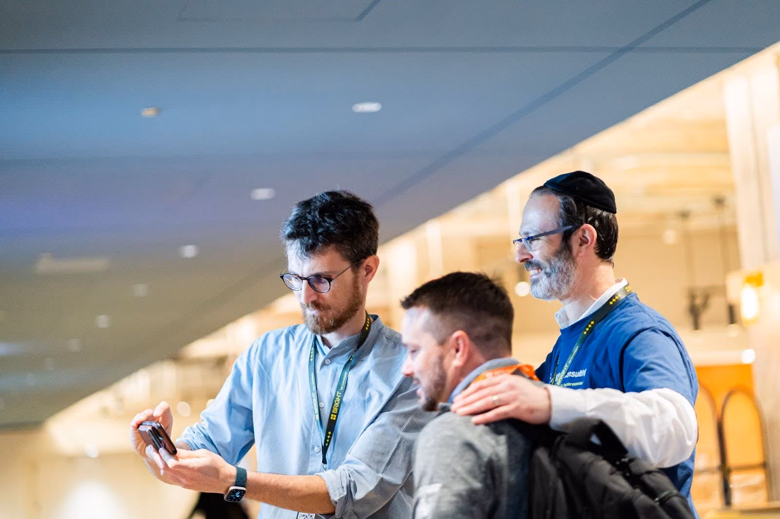 Three men, two wearing lanyards, one taking a selfie with a smartphone, indoors at an event.