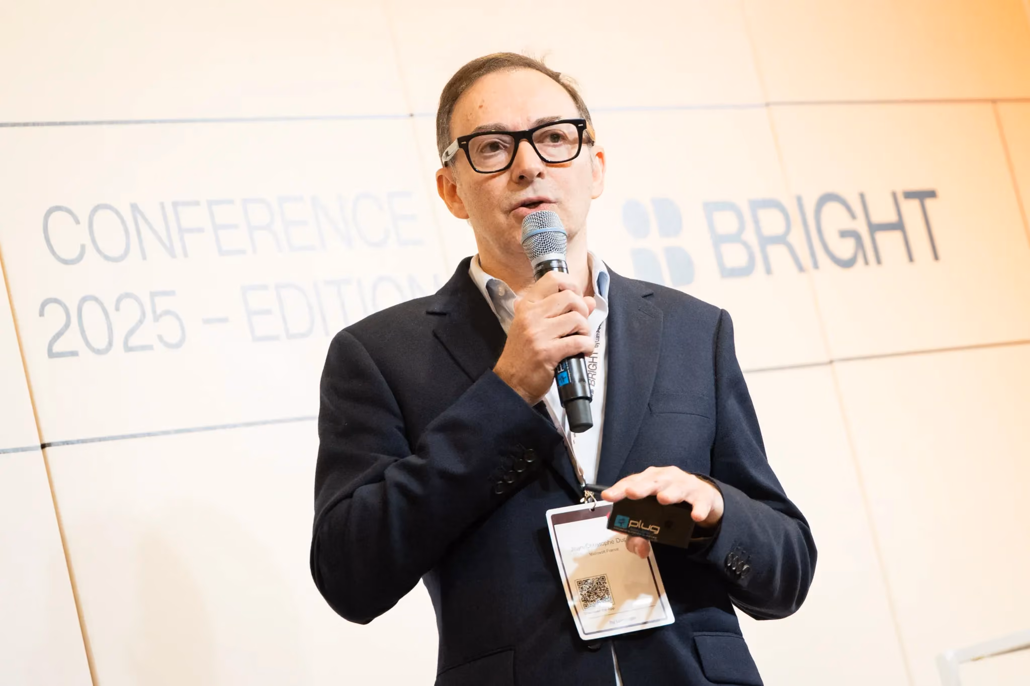 Man in glasses and suit speaking into a microphone at a conference with BRIGHT logo in background.