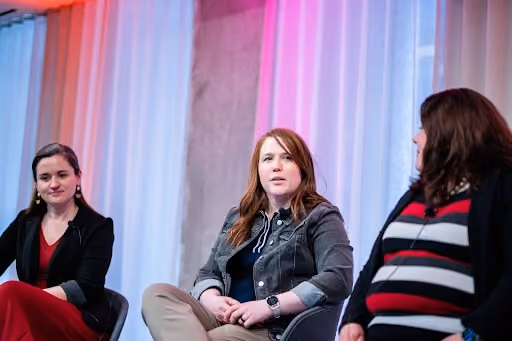 Three women seated and engaged in conversation against a backdrop of white curtains.