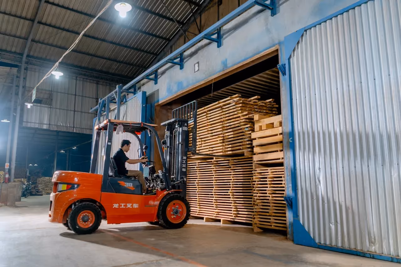Worker driving orange forklift stacking wooden planks inside a spacious warehouse.