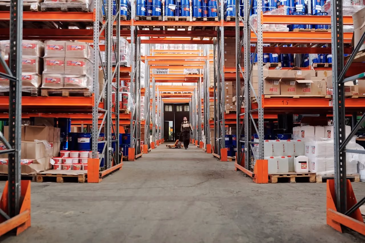 Worker walking down wide aisle between tall metal shelves stocked with pallets in a warehouse.