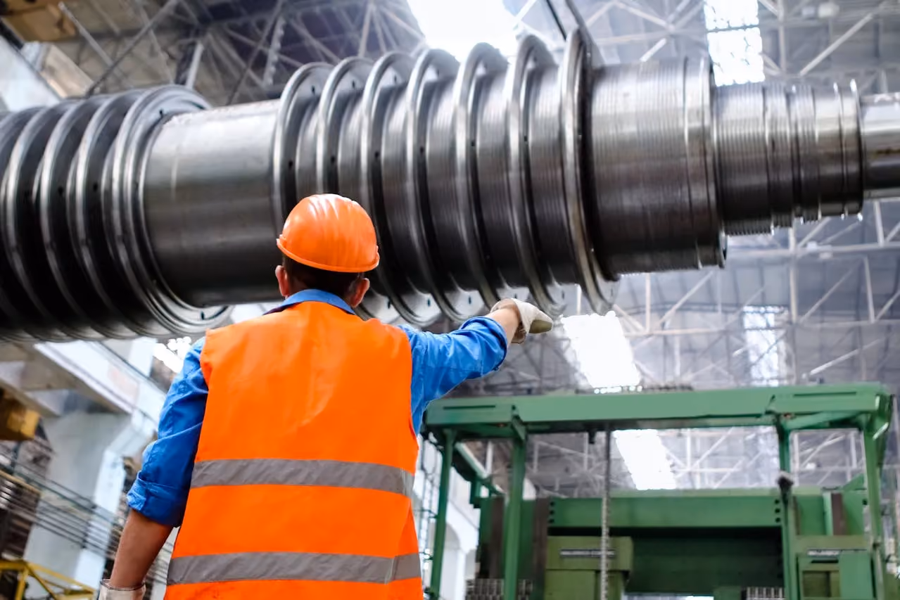 Worker in orange vest and hard hat pointing at large industrial metal screw inside factory.