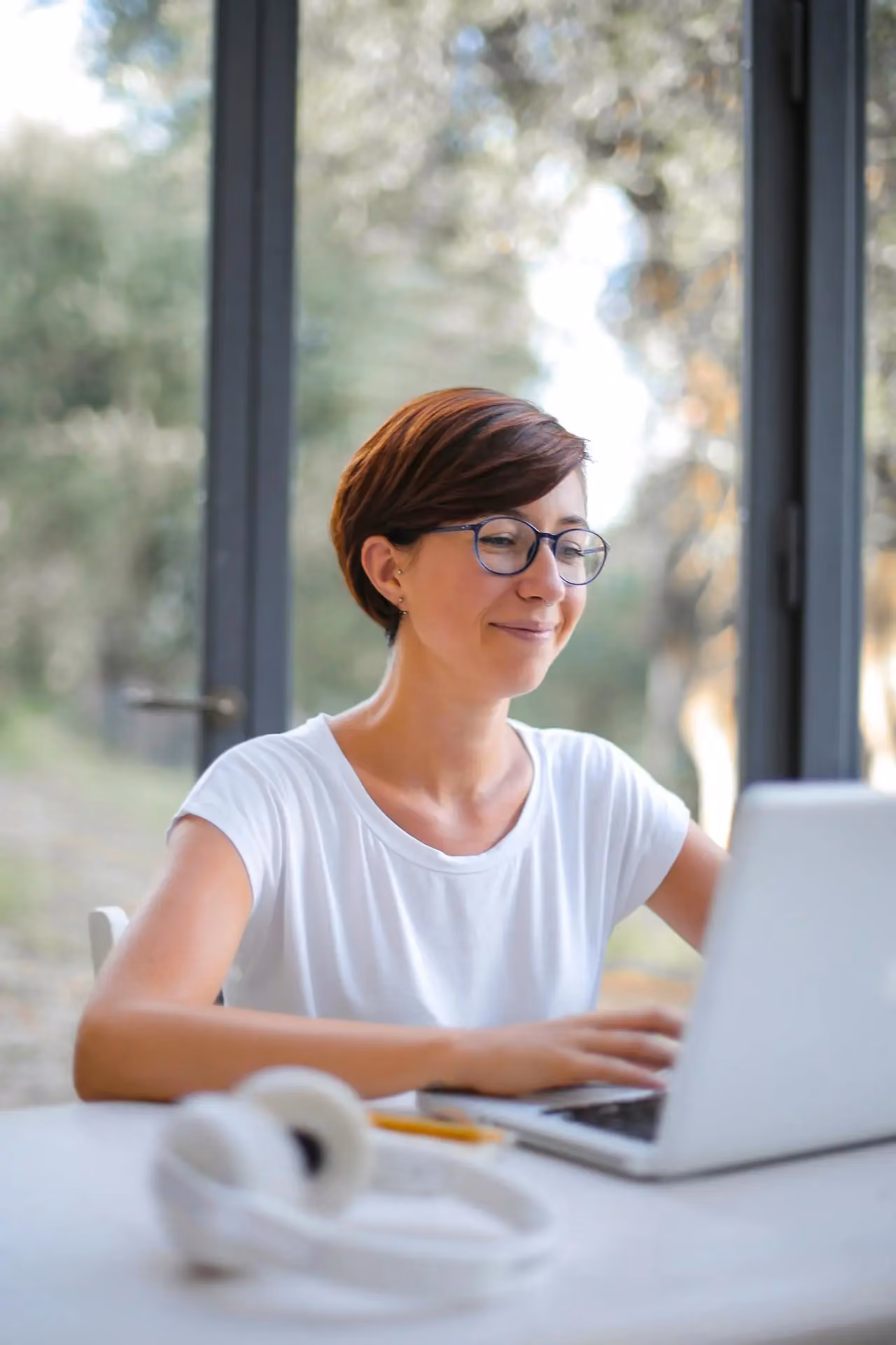 Woman with short hair and glasses working on a laptop at a white table near large windows.