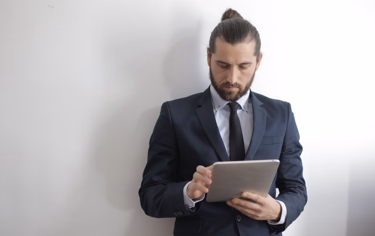 Man in a suit looking intently at a tablet he holds with both hands against a white wall.