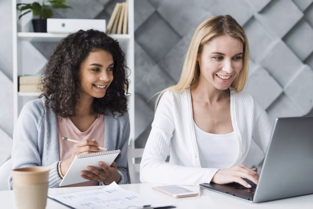 Two women smiling and working together at a desk with laptop, notebook, and papers.