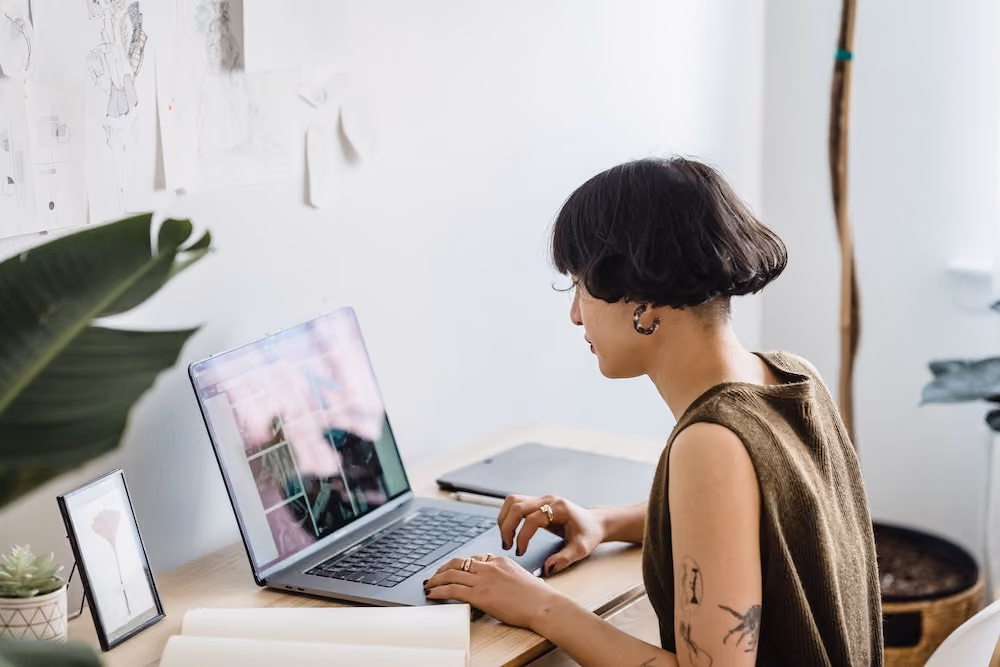 Person with short hair and tattoos working on a laptop at a desk with plants and drawings.