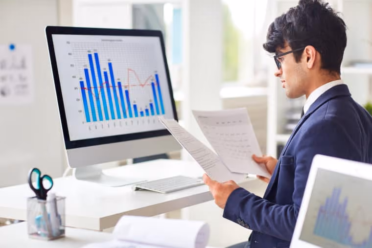Man in glasses and suit reviewing papers at desk with bar chart on computer screen.
