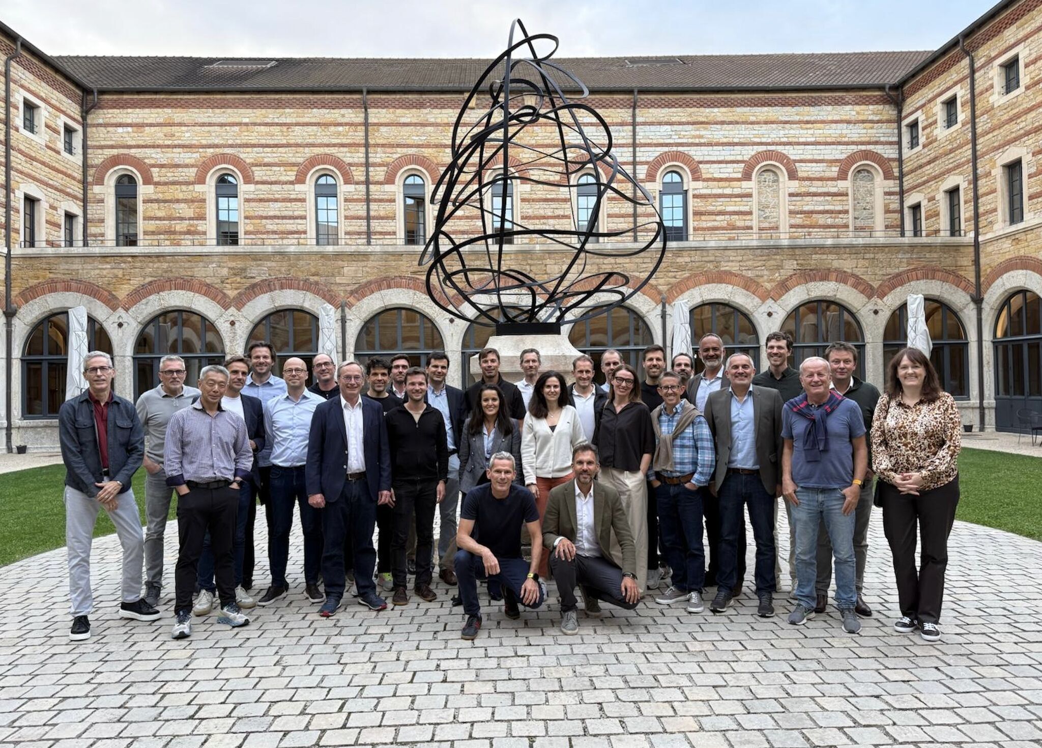 Group of 28 people posing in courtyard with brick building and abstract black sculpture behind them.