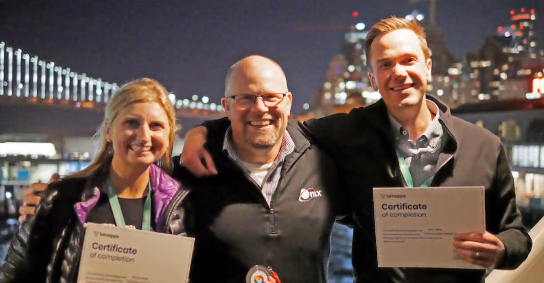 Three people smiling at night, two holding certificates of completion in front of a city bridge.
