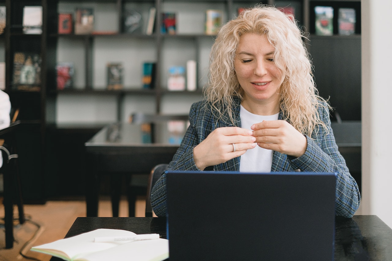Woman with curly blonde hair sitting at a table using a laptop, smiling and gesturing with hands.
