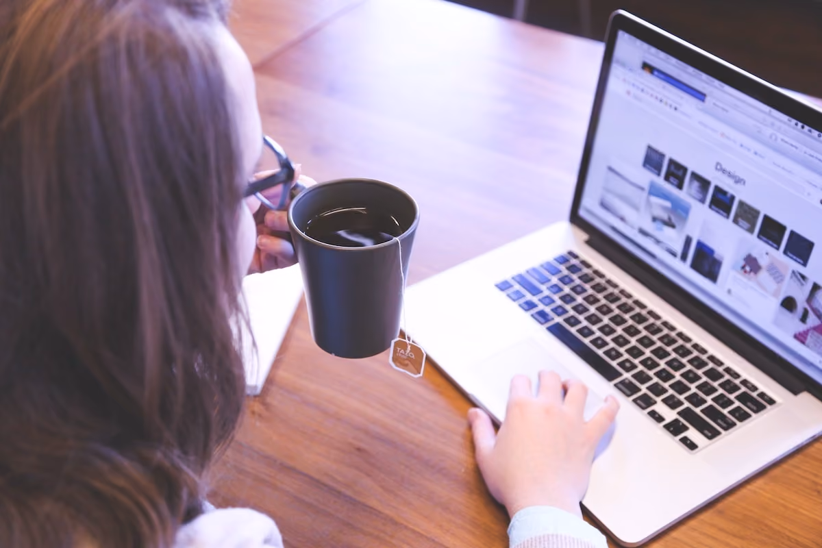 Person holding a black mug with tea while using a laptop displaying a design webpage.