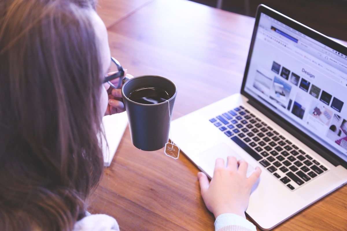 Person holding a black mug with tea while using a laptop displaying a design webpage.