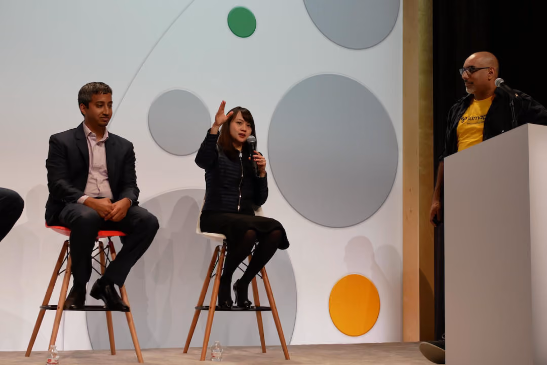 Two people sitting on stools, one speaking into a microphone, while a third person stands at a podium.