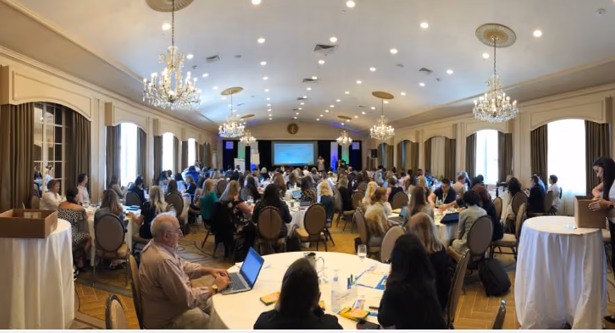 Large group attending a conference in an elegant ballroom with chandeliers and a presenter at the front.