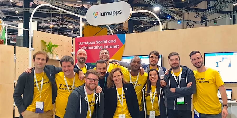 Group of LumApps team members posing together at a trade show booth with LumApps signage behind them.