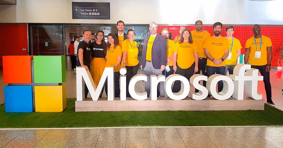 Group of people wearing LumApps T-shirts posing behind large Microsoft sign with colorful logo blocks.