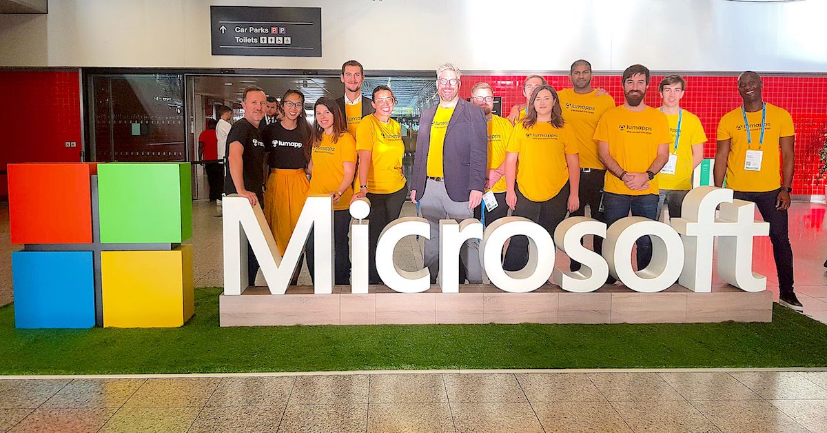 Group of people wearing LumApps T-shirts posing behind large Microsoft sign with colorful logo blocks.