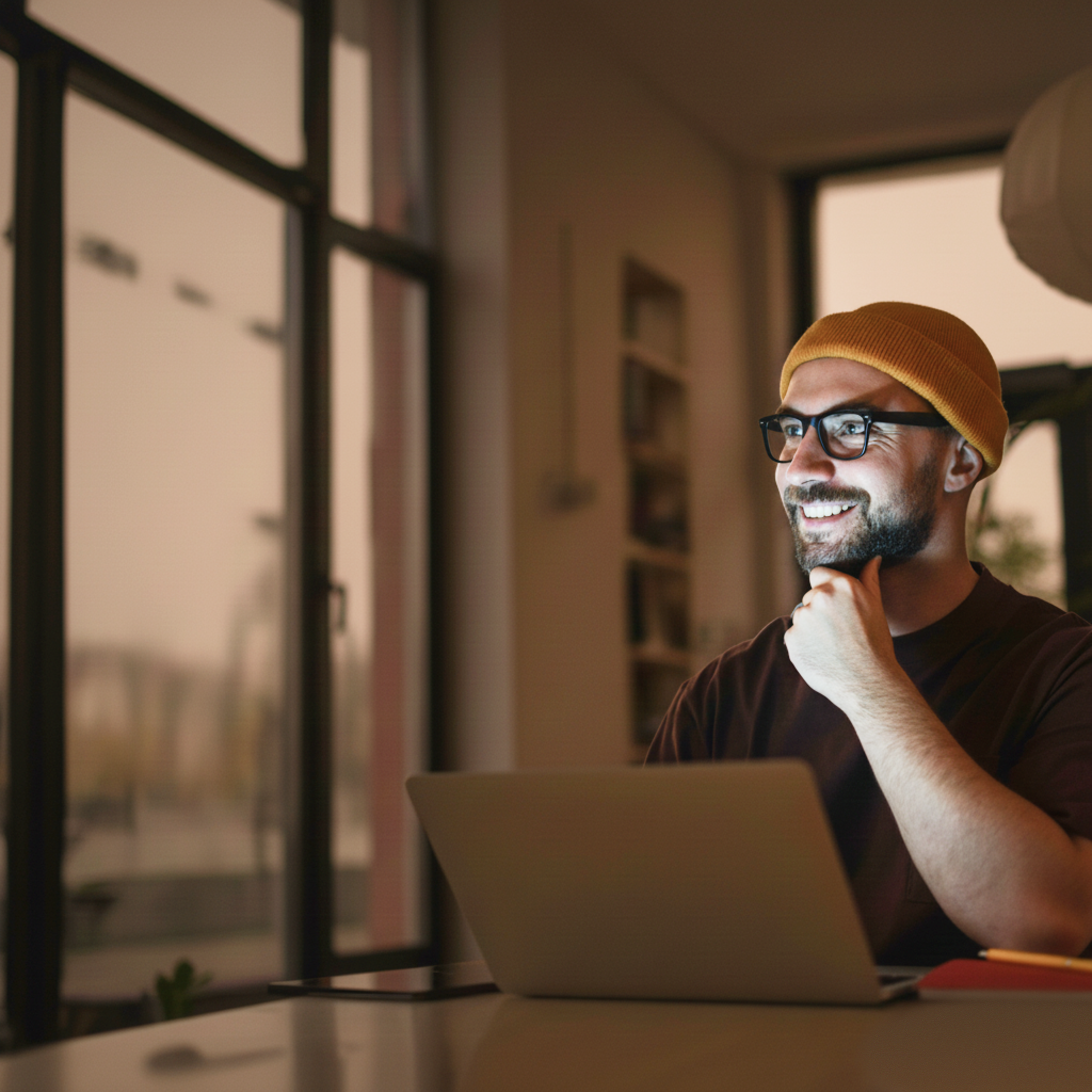 Smiling man with glasses and yellow beanie working on laptop by window at dusk.