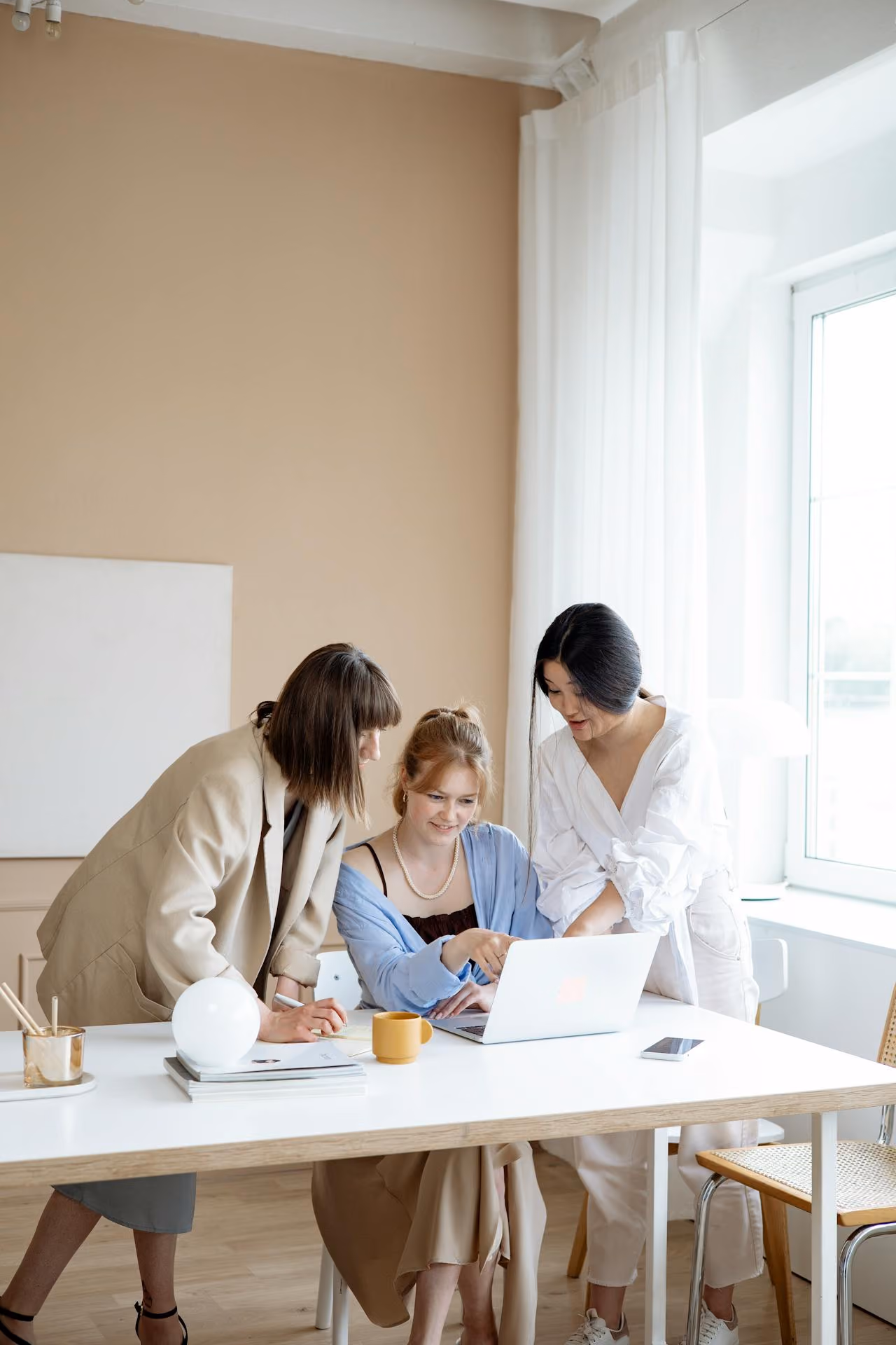 Three women collaborating around a laptop in a bright office with white walls and natural light.