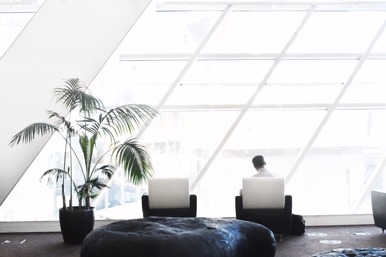 Man sitting on a chair facing large slanted windows in a bright modern room with plants and cushions.