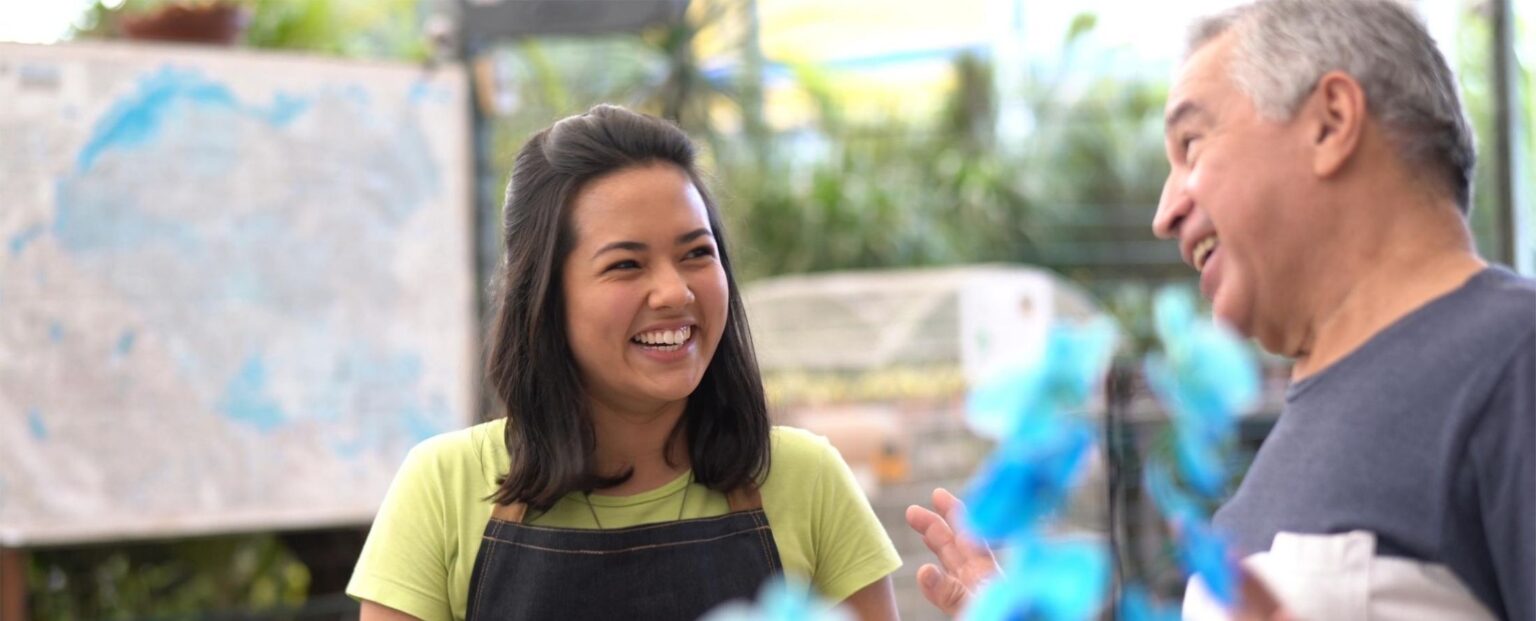 A woman wearing an apron smiles while engaging in warm interpersonal communication with an older man in a bright, indoor setting with plants in the background. Beekeeper