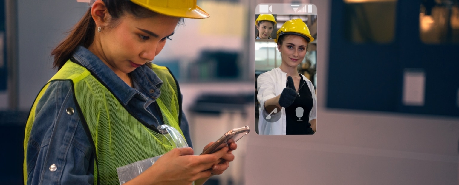 A woman in a safety vest and hard hat uses her phone in a factory, video calling another woman in similar safety gear who is giving a thumbs up. Beekeeper