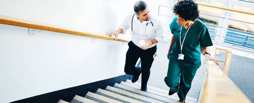 Two healthcare professionals, one in a white coat and one in green scrubs, walk up a staircase while discussing the importance of communication in healthcare within a medical facility. Beekeeper