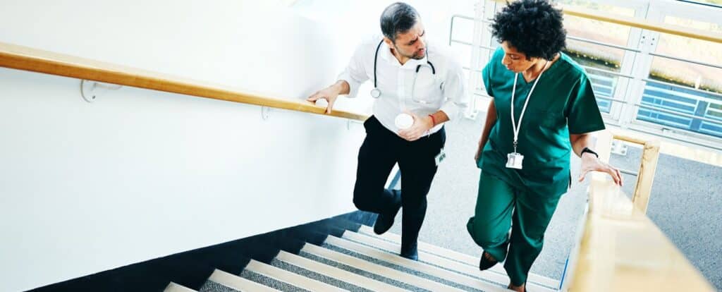 Two healthcare professionals, one in a white coat and one in green scrubs, walk up a staircase while discussing the importance of communication in healthcare within a medical facility. Beekeeper