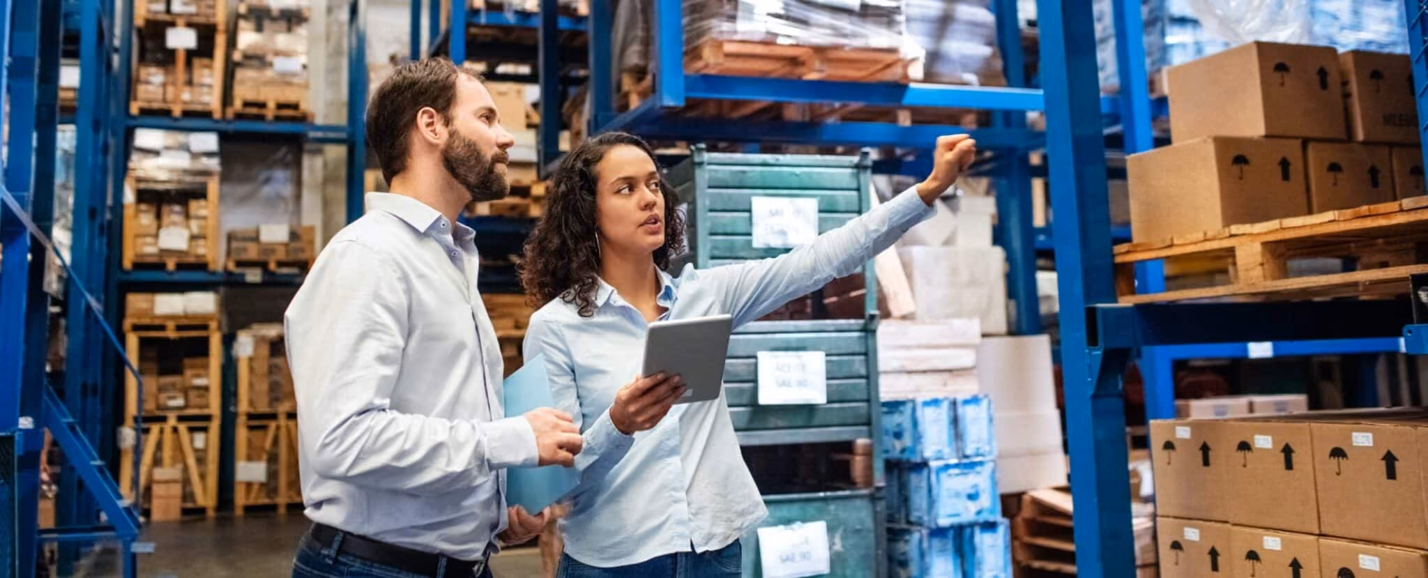 Two people in a warehouse, one holding a tablet and pointing at shelves with boxes, while the other looks on. Beekeeper