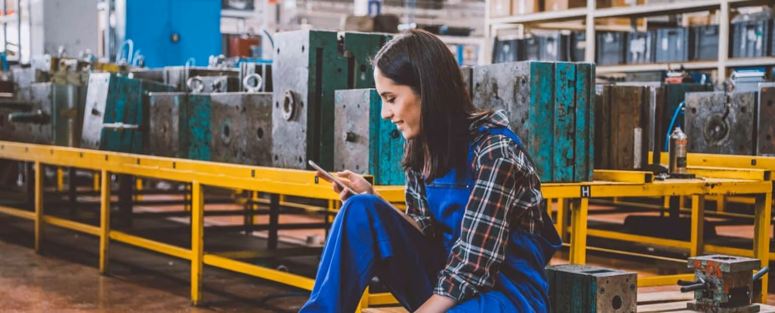 A woman in blue overalls sits on the floor of a factory, using her smartphone for mobile communication, with industrial machinery and equipment in the background. Beekeeper