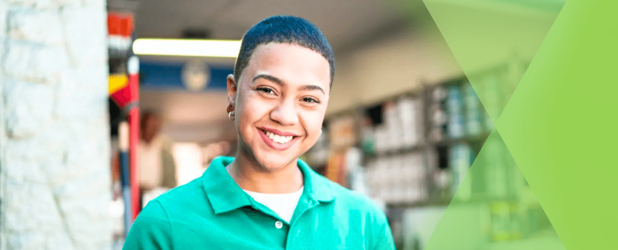 A person with short hair wearing a green collared shirt smiles indoors, with shelves and products visible in the background, suggesting an inviting setting perfect for open enrollment communication. Beekeeper