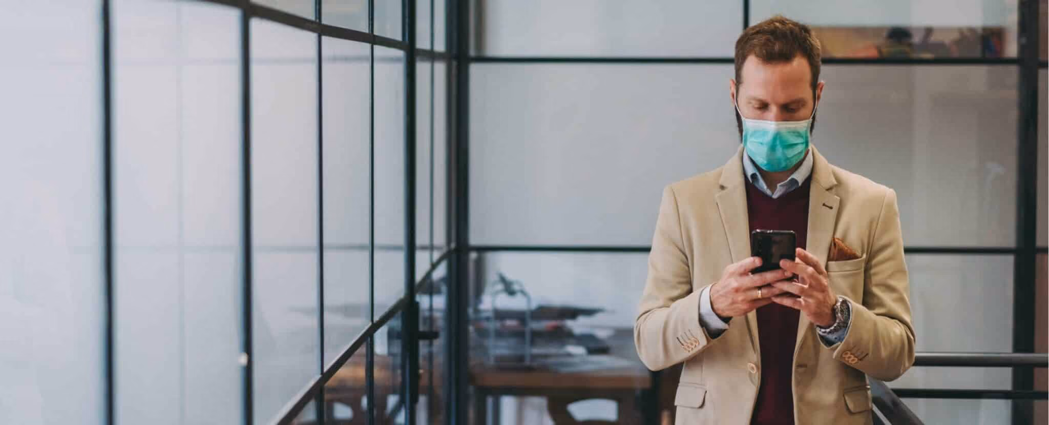 A man in a suit and face mask stands indoors, looking down at his smartphone, possibly checking business continuity tools, in a modern office setting with glass walls. Beekeeper