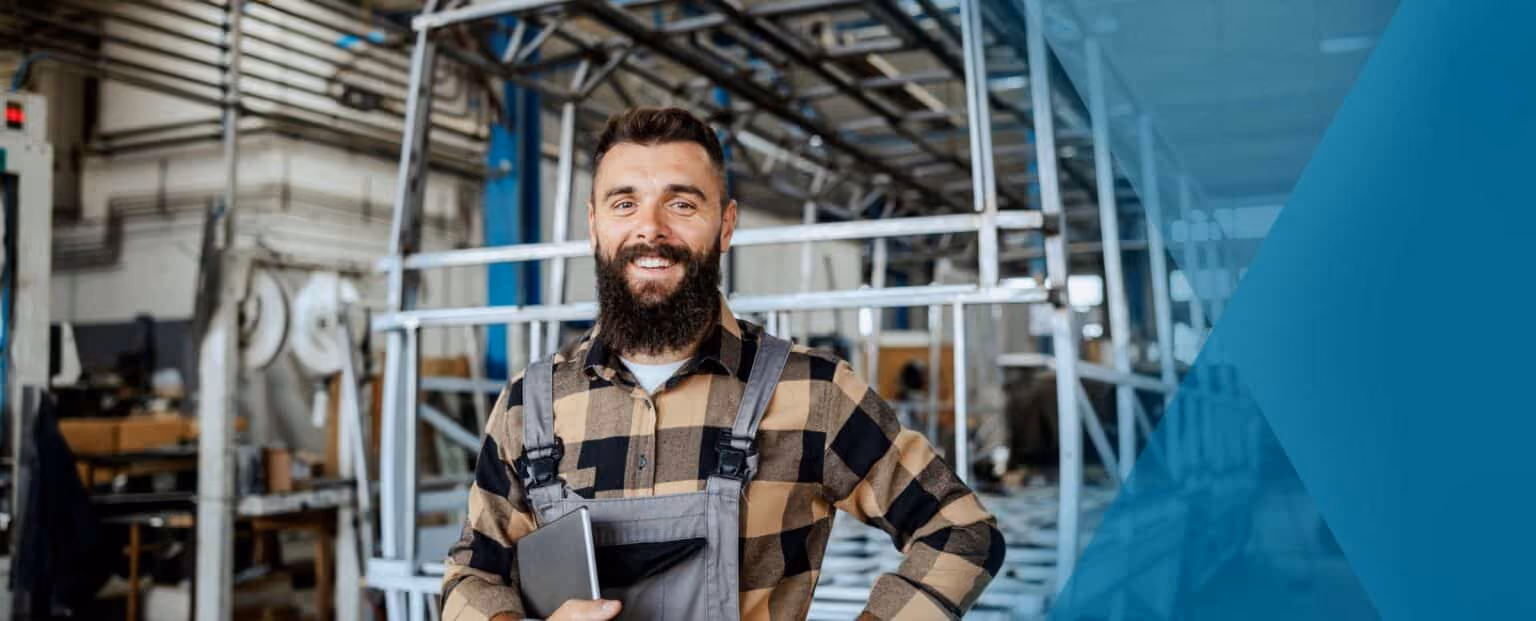 Man with a beard and plaid shirt stands in a workshop holding a tablet, reviewing employee offboarding details, with metal frames and industrial equipment in the background. Beekeeper
