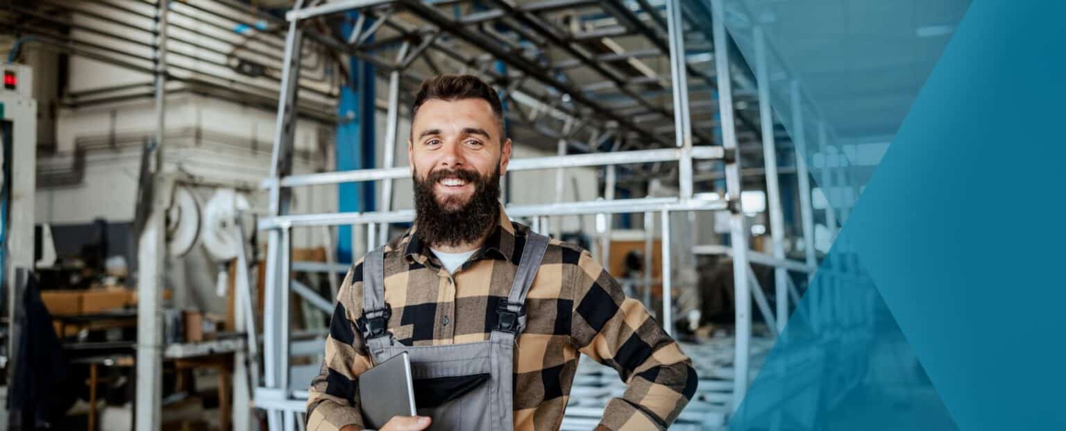 Man with a beard and plaid shirt stands in a workshop holding a tablet, reviewing employee offboarding details, with metal frames and industrial equipment in the background. Beekeeper