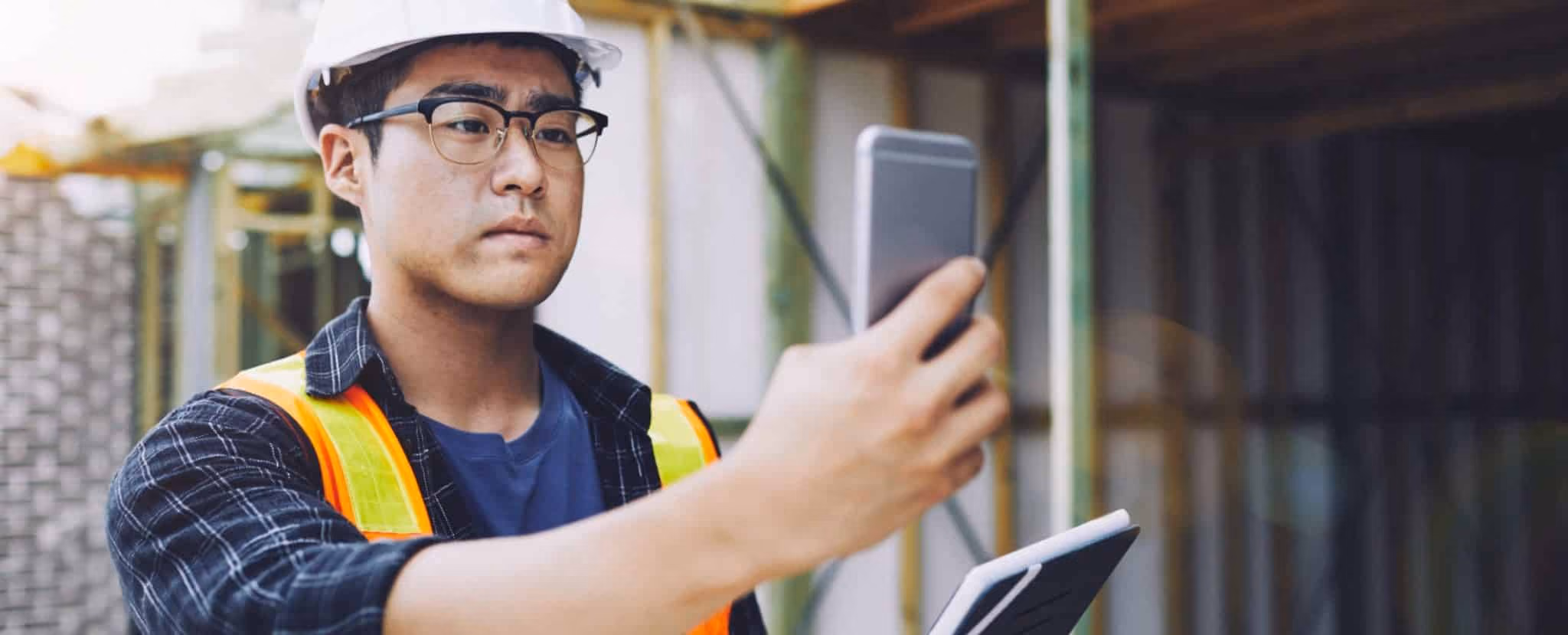 A person wearing a hard hat and safety vest uses a smartphone and holds a tablet at a construction site, emphasizing workplace safety. Beekeeper
