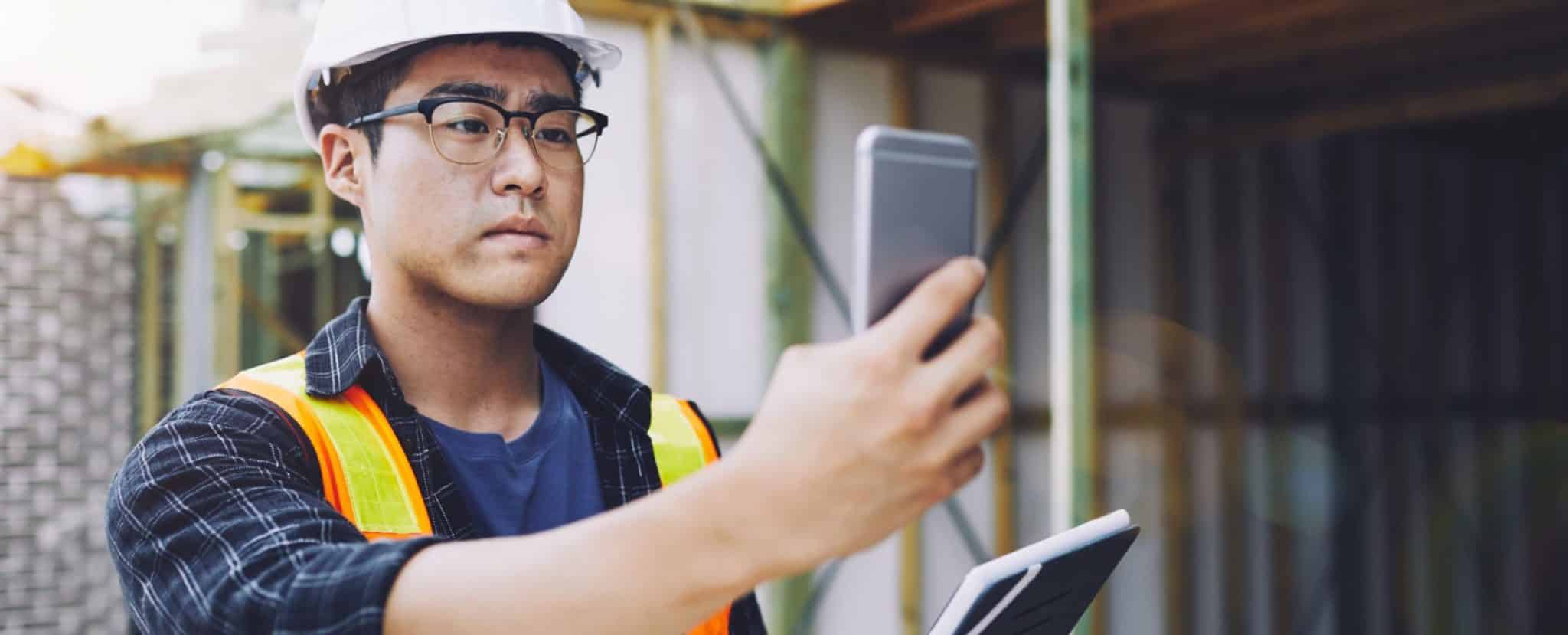 A person wearing a hard hat and safety vest uses a smartphone and holds a tablet at a construction site, emphasizing workplace safety. Beekeeper
