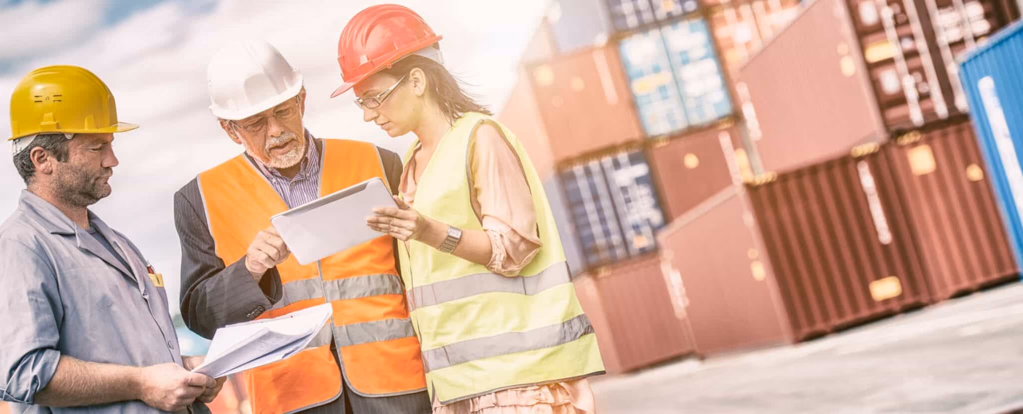 Three workers in safety gear review documents and a tablet, showcasing effective logistics communication in front of stacked shipping containers at an industrial site. Beekeeper