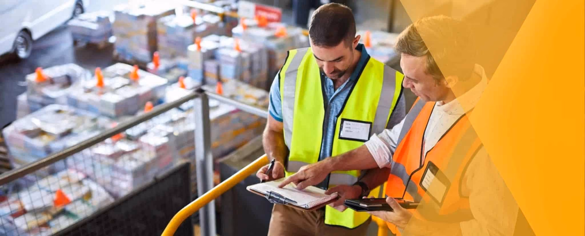 Two workers in safety vests review documents on a clipboard, using shift handover templates in a warehouse filled with stacks of packaged materials. Beekeeper