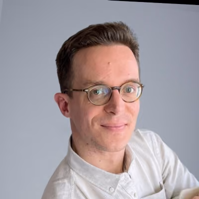 Smiling man wearing glasses and a light-colored shirt, posing against a gray background.