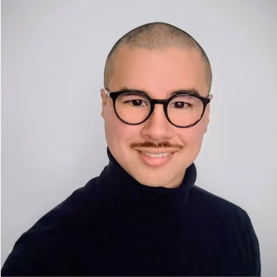 Smiling man with glasses, mustache, and shaved head wearing a black turtleneck against white background.