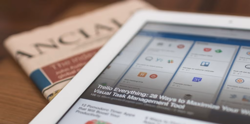 Tablet displaying Trello task management app next to a folded Financial Times newspaper on a wooden surface.